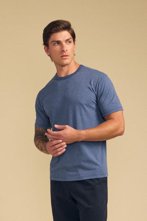A man wearing the ZANDER TEE by Velvet by Graham & Spencer in blue, paired with dark pants and brown sandals, stands against a beige background—perfect for everyday style.