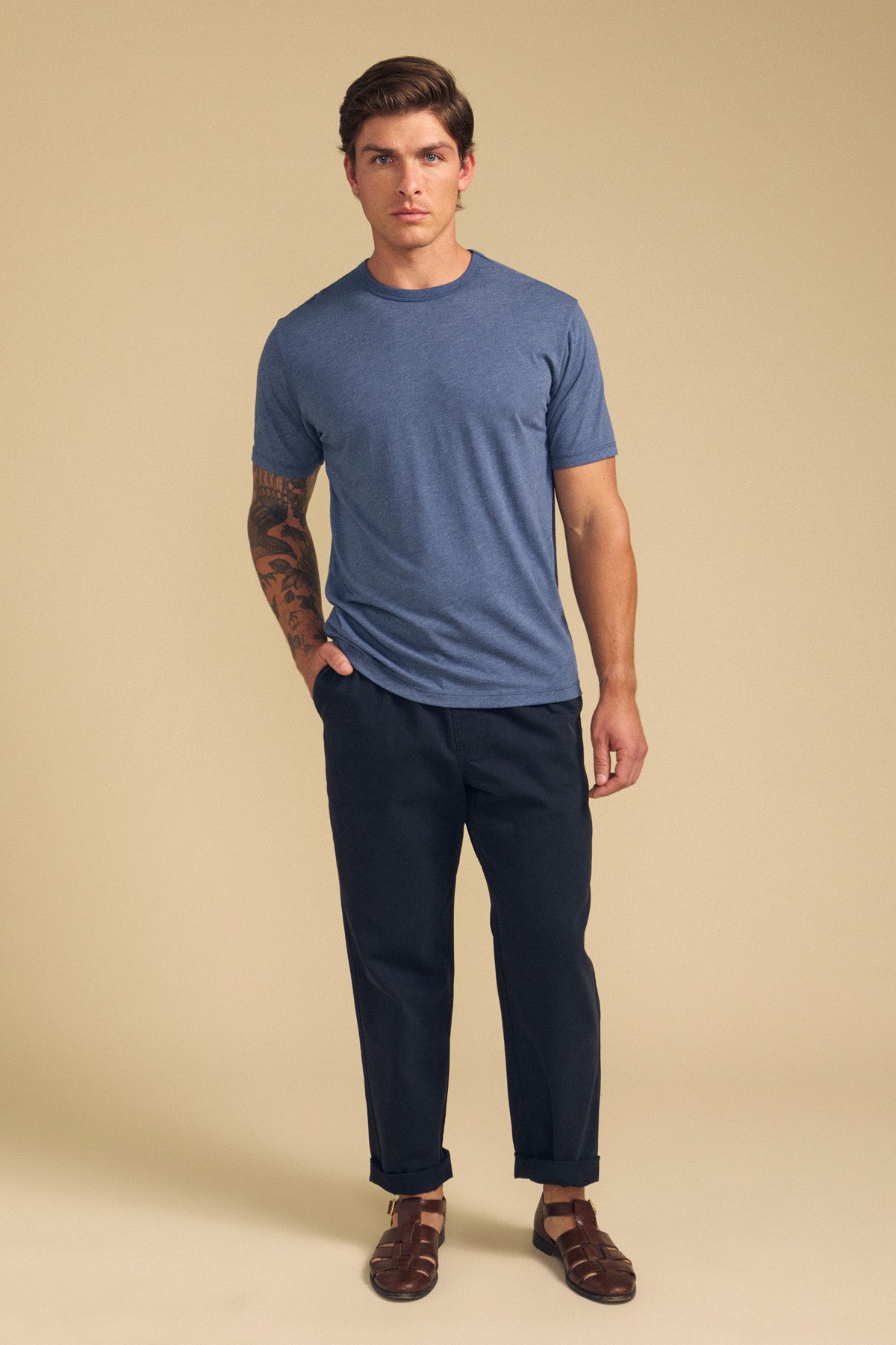A man faces forward against a beige backdrop, wearing the ZANDER TEE by Velvet by Graham & Spencer in blue, paired with dark pants and brown sandals—relaxed fit and ideal for everyday wear, with a tattoo visible on his left arm.