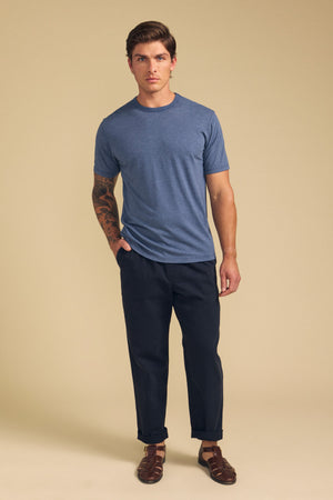 A man faces forward against a beige backdrop, wearing the ZANDER TEE by Velvet by Graham & Spencer in blue, paired with dark pants and brown sandals—relaxed fit and ideal for everyday wear, with a tattoo visible on his left arm.