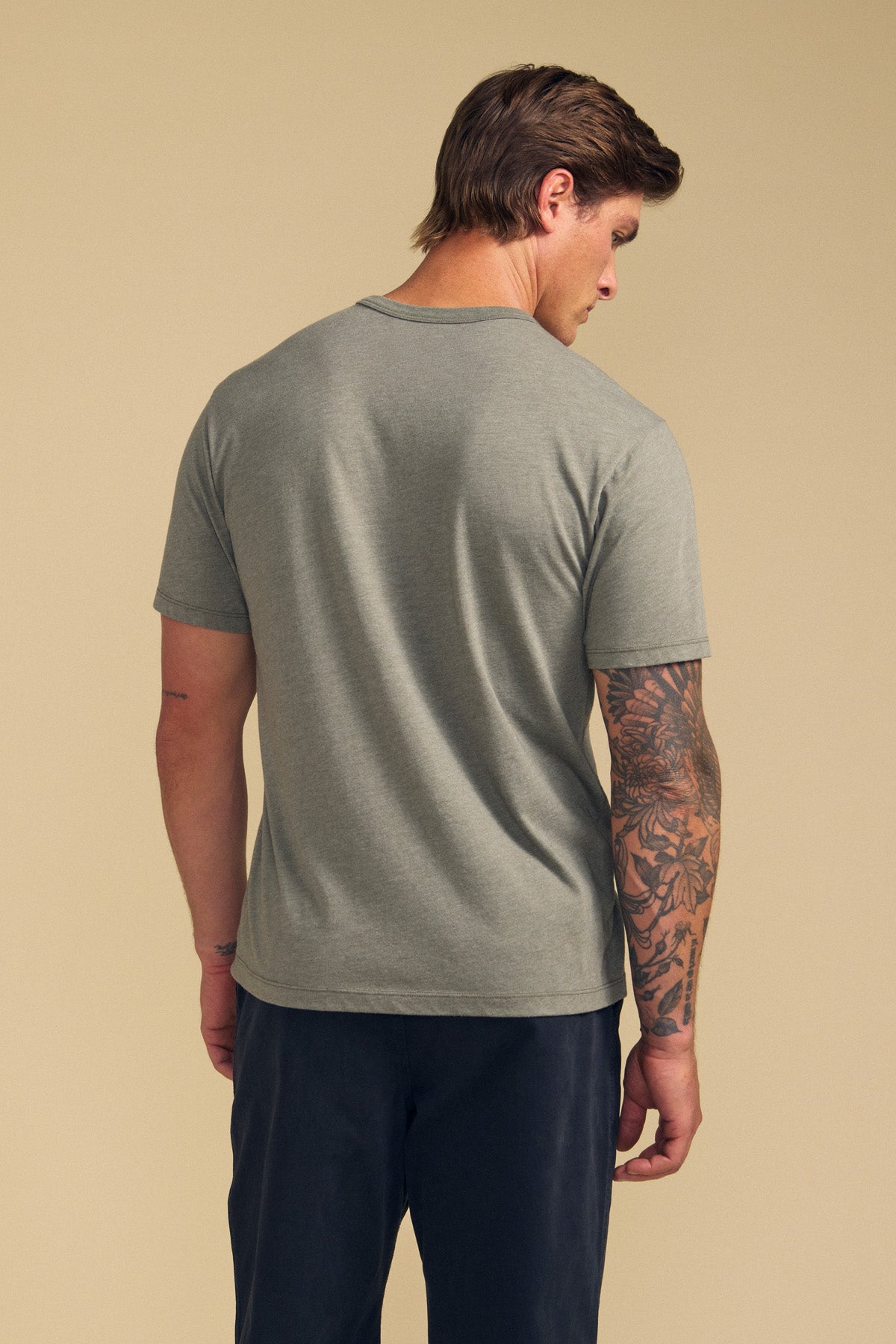 A man with short brown hair and a tattooed right arm stands with his back to the camera, wearing the ZANDER TEE by Velvet by Graham & Spencer in gray—a relaxed fit cotton-modal shirt, paired with dark pants against a beige background.