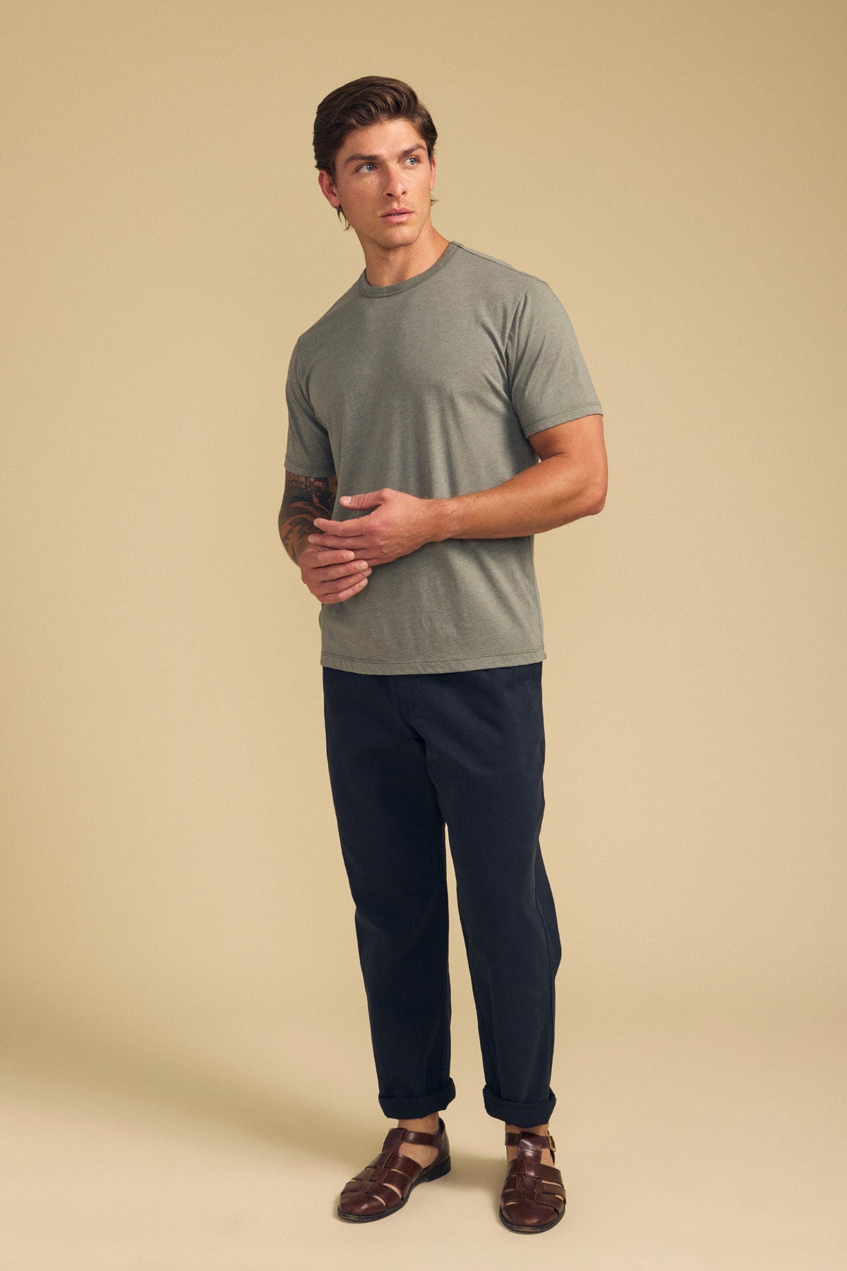A man stands before a beige background wearing the ZANDER TEE by Velvet by Graham & Spencer—a relaxed fit, grey cotton-modal blend t-shirt—paired with dark pants and brown sandals, hands clasped and gaze to the side; ideal for everyday style.-41702129631425