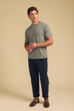A man stands before a beige background wearing the ZANDER TEE by Velvet by Graham & Spencer—a relaxed fit, grey cotton-modal blend t-shirt—paired with dark pants and brown sandals, hands clasped and gaze to the side; ideal for everyday style.