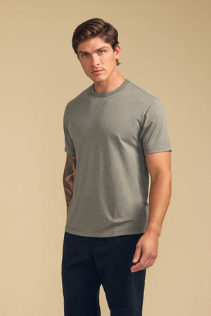 A man with short brown hair and a tattoo on his right forearm wears the ZANDER TEE by Velvet by Graham & Spencer in light gray along with dark pants, standing against a beige background.