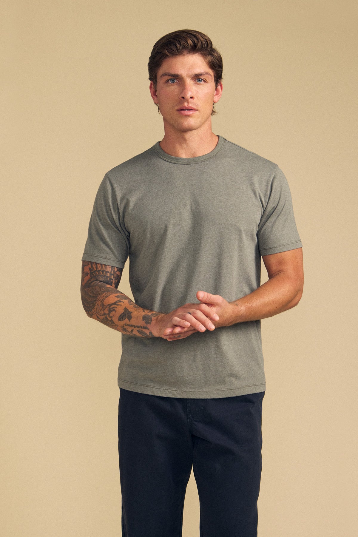 A man with a tattooed right arm stands against a beige background, wearing the Velvet by Graham & Spencer ZANDER TEE—a relaxed fit grey cotton-modal blend short-sleeve top—and dark pants, ideal for everyday style.