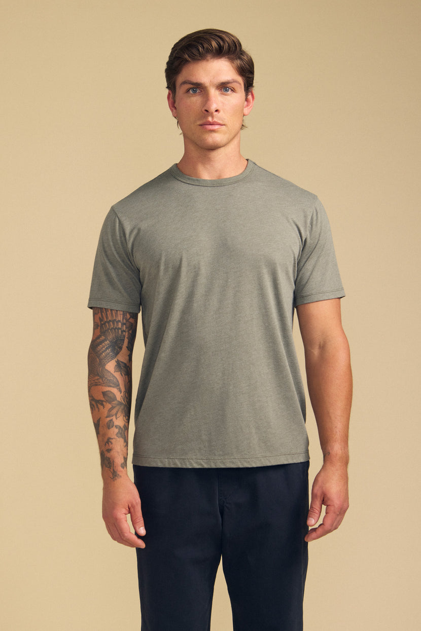 A man with a tattoo on his left forearm and a neutral expression stands facing forward against a beige background, wearing the Velvet by Graham & Spencer ZANDER TEE in soft gray cotton-modal and dark pants.