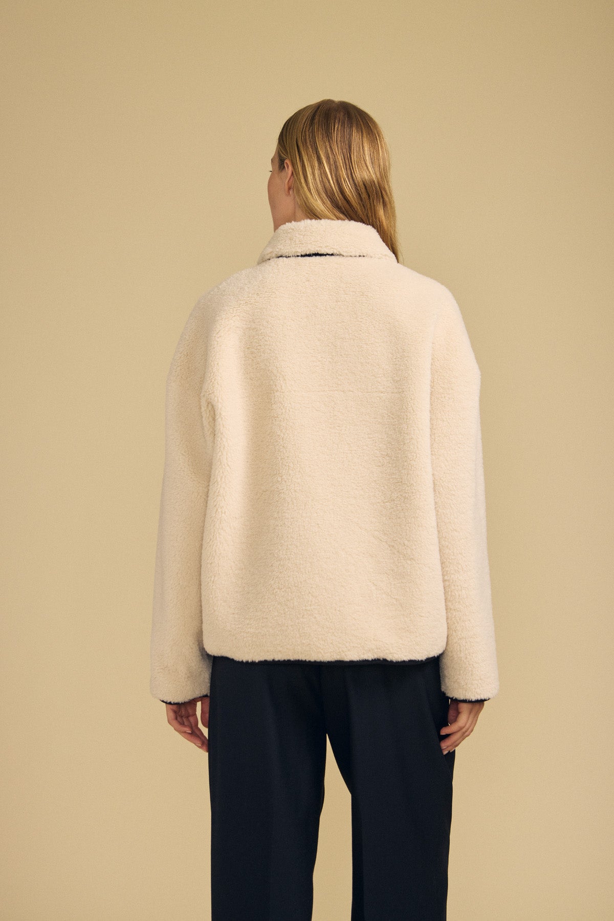 A person with long blonde hair faces away, wearing the cream-colored ANIKA TEDDY JACKET by Velvet by Graham & Spencer paired with black pants against a plain beige background—the perfect look for cold-weather layering.-41935330181313