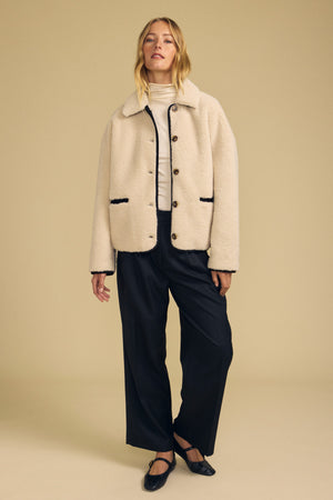 A person stands against a plain beige background wearing the ANIKA TEDDY JACKET by Velvet by Graham & Spencer layered over a white turtleneck, paired with black wide-leg pants and black shoes—an effortlessly stylish look, perfect for cold-weather layering.
