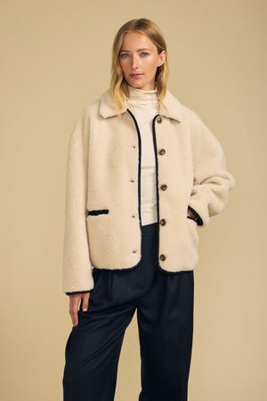 A woman models the ANIKA TEDDY JACKET by Velvet by Graham & Spencer—its cream-colored faux sherpa texture is layered over a white turtleneck and loose black pants, creating a cozy, cold-weather look against a beige background.