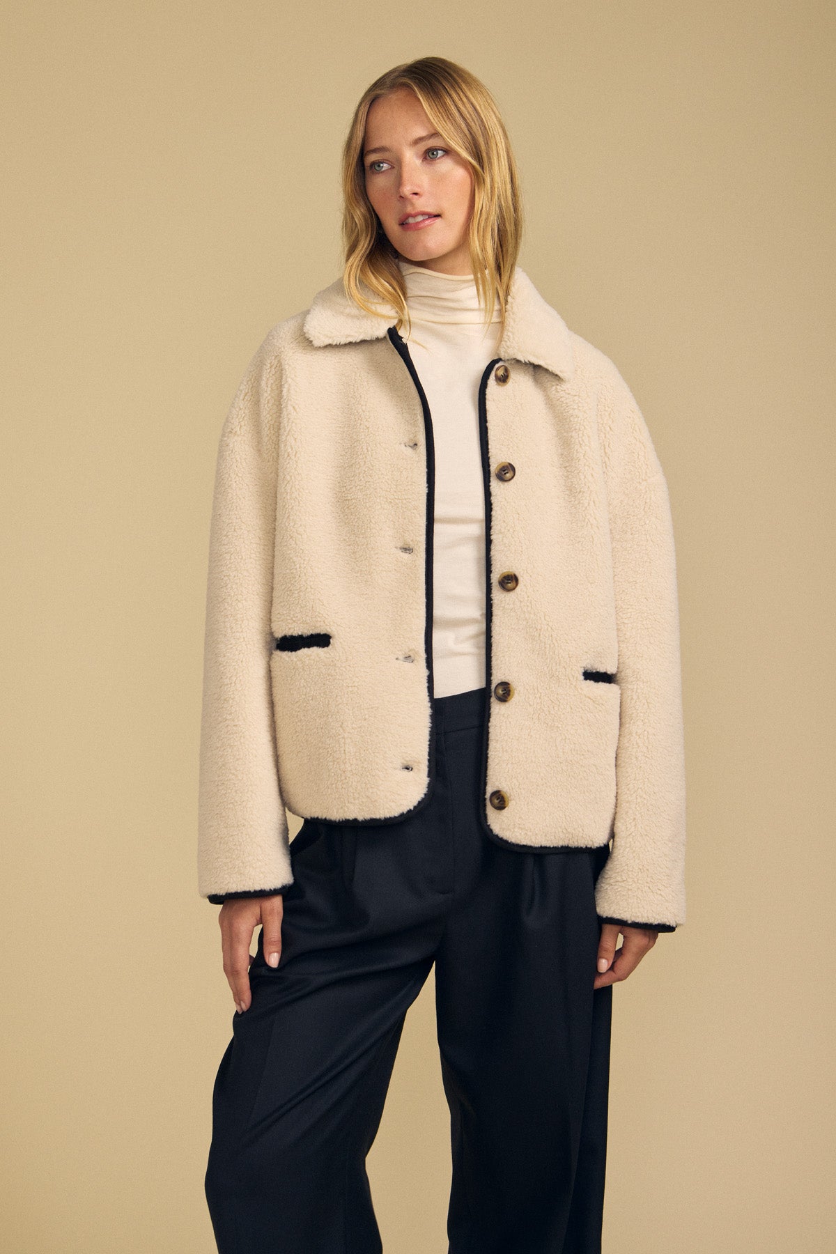 Clad in the ANIKA TEDDY JACKET by Velvet by Graham & Spencer, a woman pairs this plush sherpa layer with a white turtleneck and black trousers against a plain beige background—a cozy option for cold-weather layering.