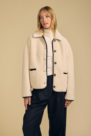 Clad in the ANIKA TEDDY JACKET by Velvet by Graham & Spencer, a woman pairs this plush sherpa layer with a white turtleneck and black trousers against a plain beige background—a cozy option for cold-weather layering.