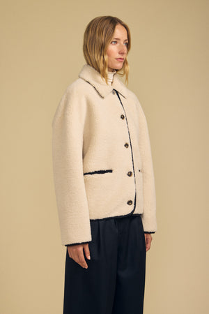 A woman stands against a plain background wearing the ANIKA TEDDY JACKET by Velvet by Graham & Spencer—a cream-colored, button-up plush sherpa jacket with black trim—paired with black pants for perfect cold-weather layering.