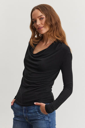 A person with wavy hair stands against a plain background, wearing the PATTY TEE by Velvet by Graham & Spencer in black and blue jeans, with one hand tucked into a pocket.