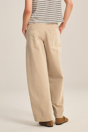 A person wearing a striped short-sleeve shirt, brown shoes, and Velvet by Graham & Spencer’s TENLEY PANT—a relaxed fit cotton twill pant with back pockets—is standing facing away from the camera.