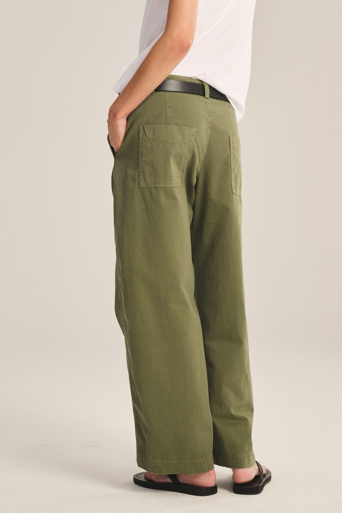 Person wearing a white t-shirt, Velvet by Graham & Spencer TENLEY PANT in relaxed fit olive green cotton twill with back patch pockets, black belt, and black flip-flops, standing with hands in pockets.-40808733245633