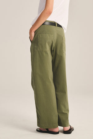 Person wearing a white t-shirt, Velvet by Graham & Spencer TENLEY PANT in relaxed fit olive green cotton twill with back patch pockets, black belt, and black flip-flops, standing with hands in pockets.