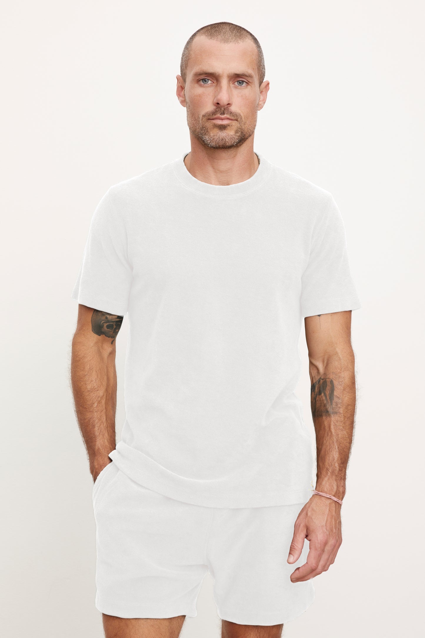 Man with light skin and shaved head wearing a white JAXON CREW t-shirt by Velvet by Graham & Spencer and shorts, standing against a plain background, with visible tattoos on his arms.-36891087732929