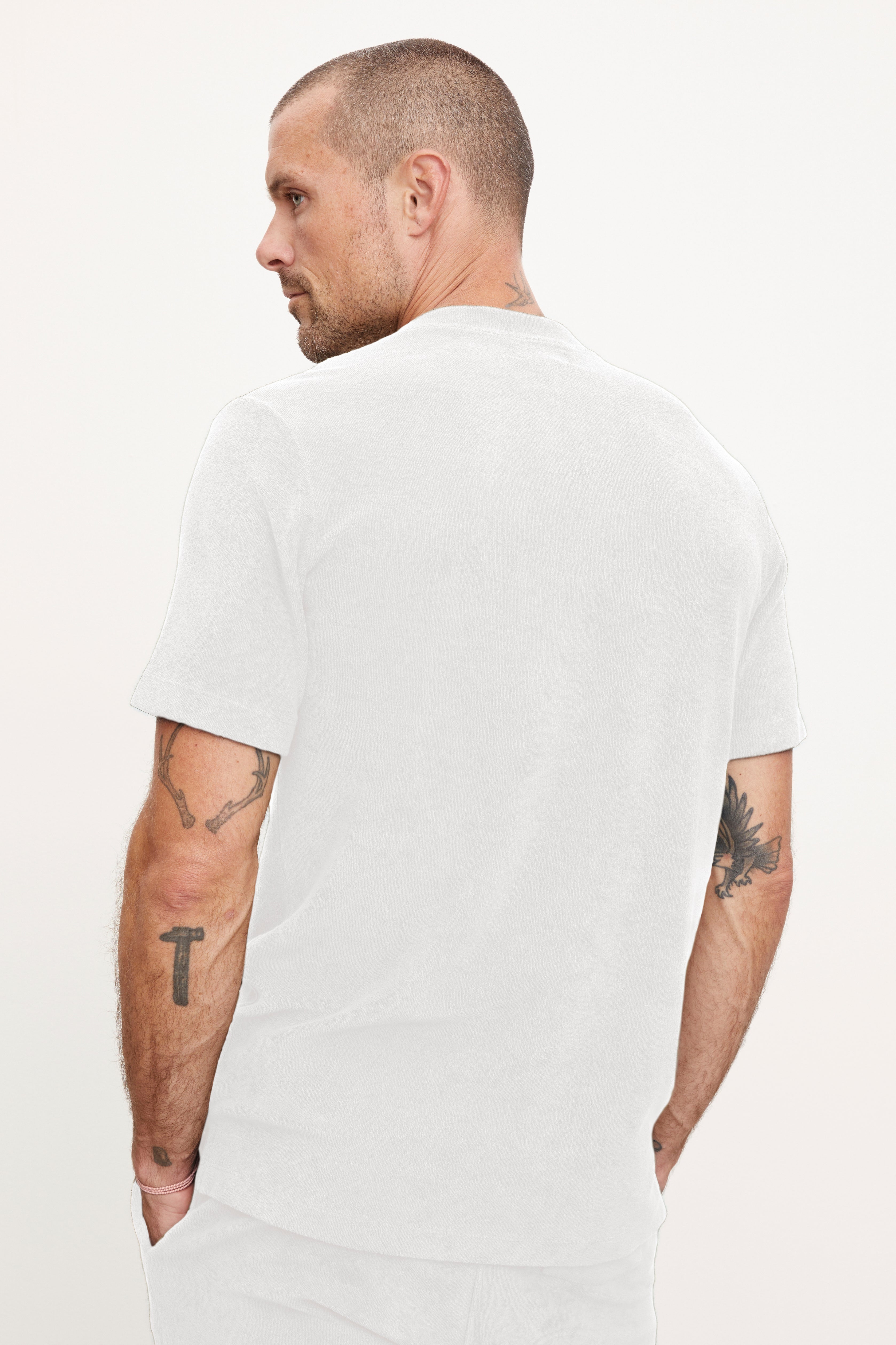   A man viewed from behind, wearing a Velvet by Graham & Spencer JAXON CREW t-shirt with a crew neckline, showing tattooed arms with visible designs, including a crucifix and a bird. 