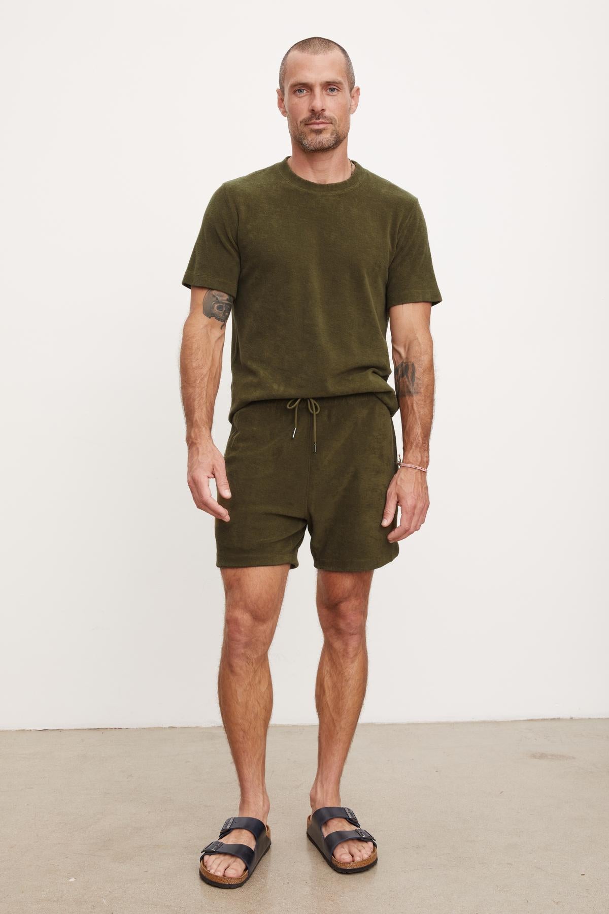 Man in a green t-shirt and Velvet by Graham & Spencer Salem shorts standing against a plain background, wearing black sandals.-36753587667137