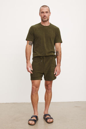 Man in a green t-shirt and Velvet by Graham & Spencer Salem shorts standing against a plain background, wearing black sandals.