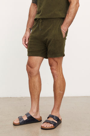 Man standing in a studio wearing green Velvet by Graham & Spencer Salem shorts and black sandals, focusing on his lower half.
