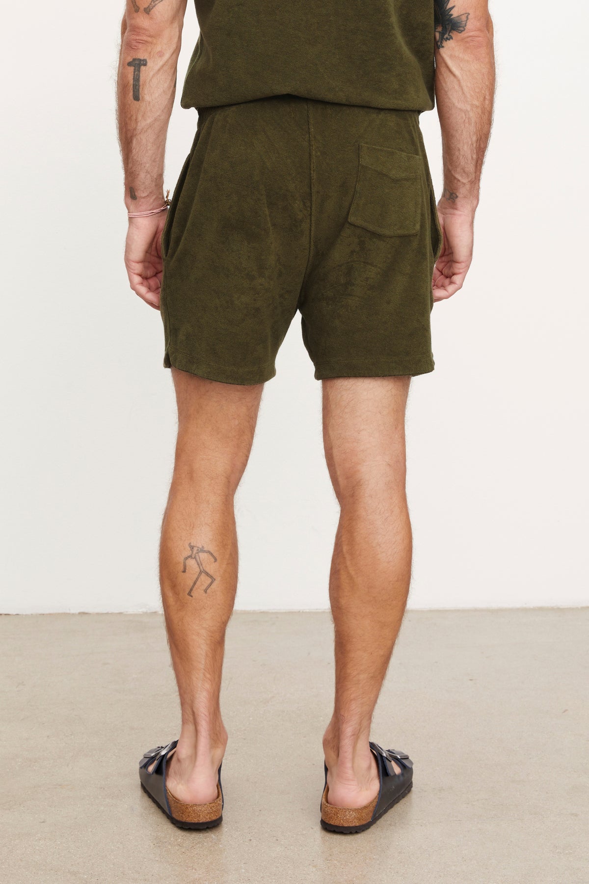   Rear view of a man wearing Velvet by Graham & Spencer's Salem shorts and brown loafers, standing with hands slightly behind him, highlighting visible tattoos on his arms and legs. 