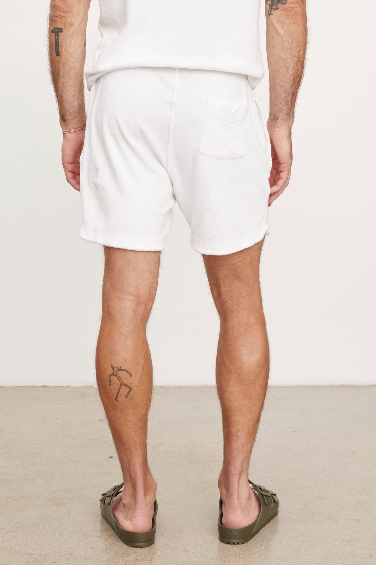   Man standing in white cotton terry cloth SALEM SHORTS by Velvet by Graham & Spencer with a back view, showcasing a tattoo on his left calf and wearing green flip-flops. 