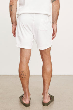 Man standing in white cotton terry cloth SALEM SHORTS by Velvet by Graham & Spencer with a back view, showcasing a tattoo on his left calf and wearing green flip-flops.
