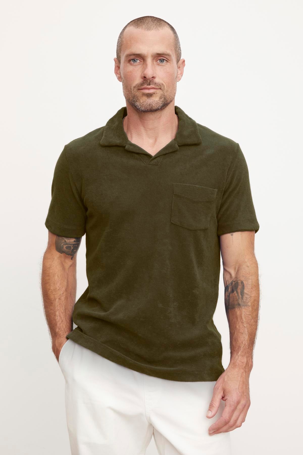 A man with short hair and tattoos on his arms, wearing a Velvet by Graham & Spencer SERGEY POLO polo shirt in dark green with short sleeves and white pants, standing against a light background.-36890717847745