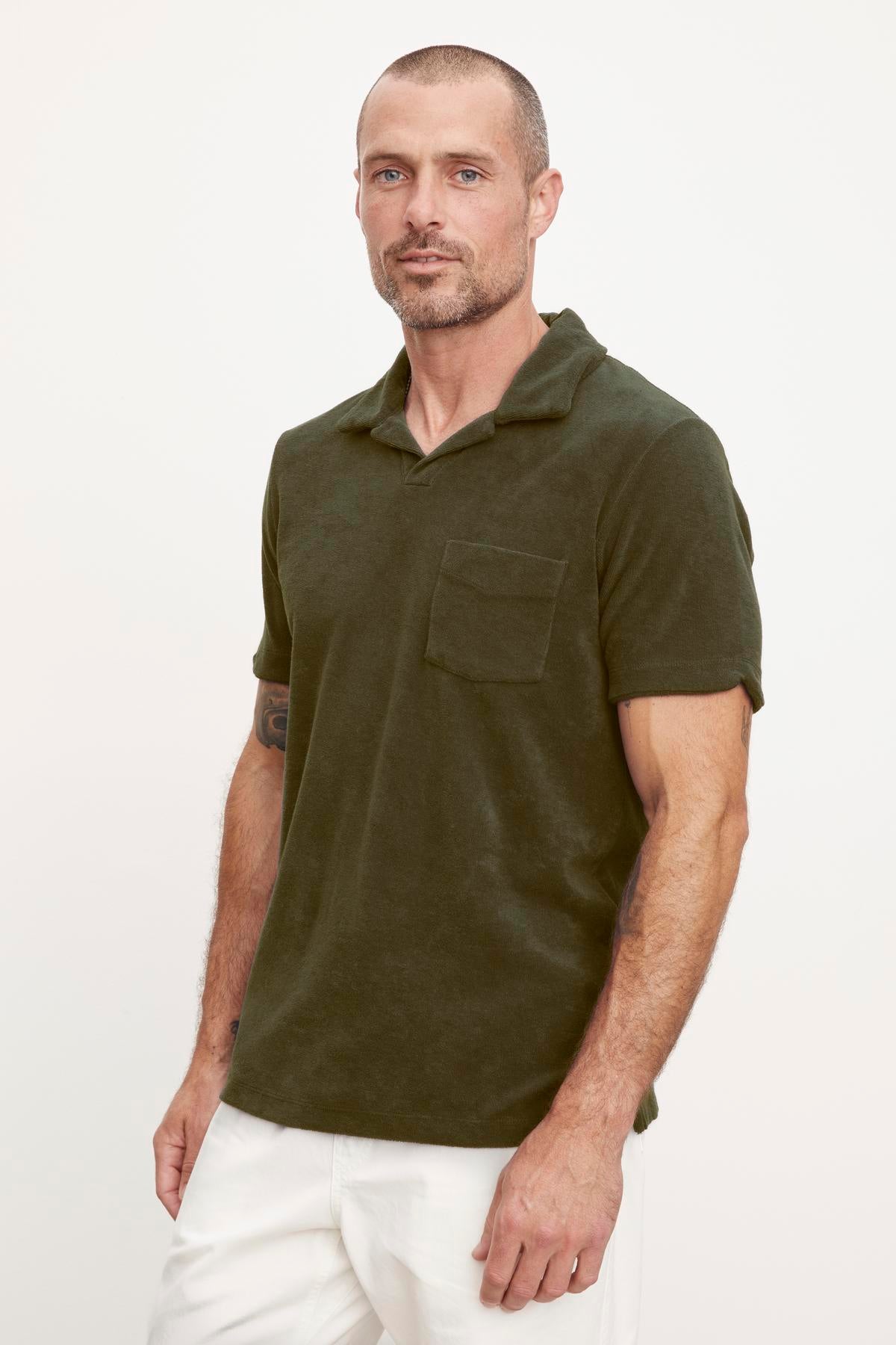 A man with a beard, wearing a dark green Sergey Polo shirt by Velvet by Graham & Spencer and white pants, standing against a plain background.-36890717880513