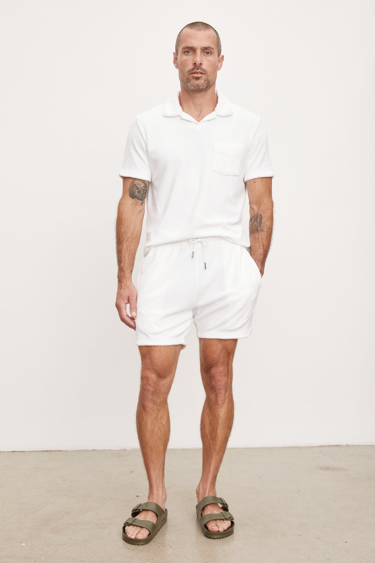 A man wearing a white polo shirt and Velvet by Graham & Spencer's SALEM SHORT drawstring elastic waist lounging shorts stands against a plain background, sporting sandals and visible tattoos on his arms.-36943667822785