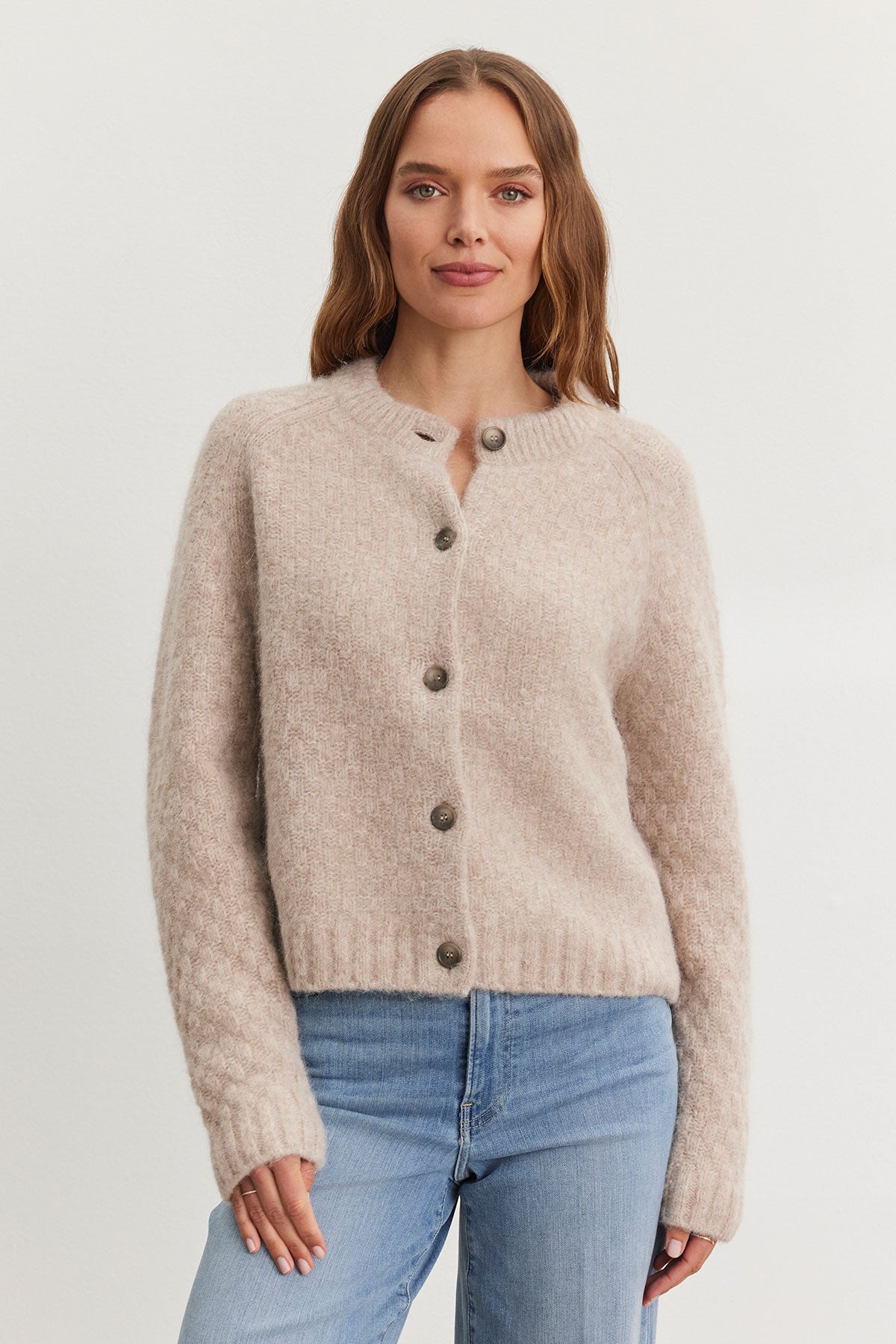   A person wearing the CHANTAL ALPACA CARDIGAN from Velvet by Graham & Spencer, featuring a beige alpaca-blend with a five-button front and ribbed trims, paired with blue jeans, stands against a plain backdrop. 