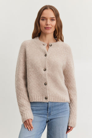 A person wearing the CHANTAL ALPACA CARDIGAN from Velvet by Graham & Spencer, featuring a beige alpaca-blend with a five-button front and ribbed trims, paired with blue jeans, stands against a plain backdrop.