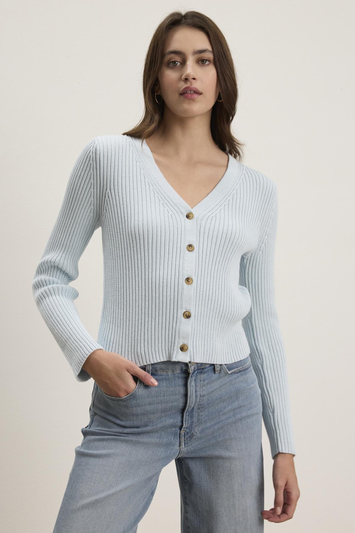   A person wearing the HYDIE CARDIGAN by Velvet by Graham & Spencer, a light blue ribbed knit cardigan with buttons, pairs it with light blue jeans against a plain backdrop. 