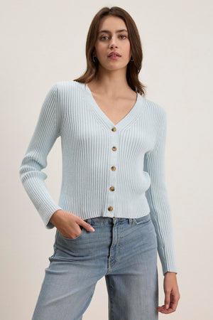 A person wearing the HYDIE CARDIGAN by Velvet by Graham & Spencer, a light blue ribbed knit cardigan with buttons, pairs it with light blue jeans against a plain backdrop.