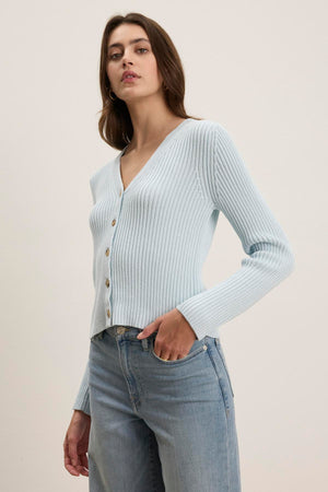 A person stands against a plain background, wearing the HYDIE CARDIGAN by Velvet by Graham & Spencer, a light blue ribbed knit with buttons, expertly paired with matching light blue jeans.