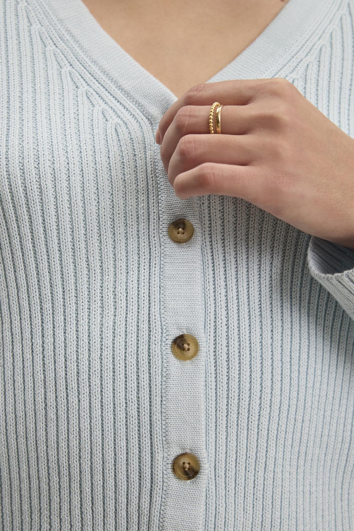   A person is shown wearing the HYDIE CARDIGAN by Velvet by Graham & Spencer, a light blue ribbed knit design with brown buttons, hand near the top button adorned with a gold ring. 