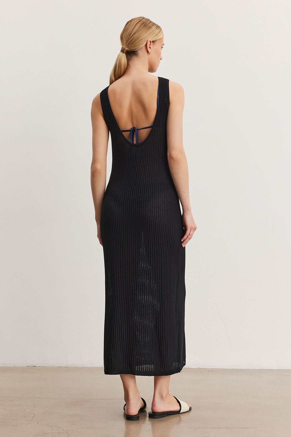 A woman stands facing away, wearing a CALLEN DRESS by Velvet by Graham & Spencer, black and ribbed with a low back and small bow detail. She has blonde hair tied in a low bun and wears black flat sandals.-36998731890881