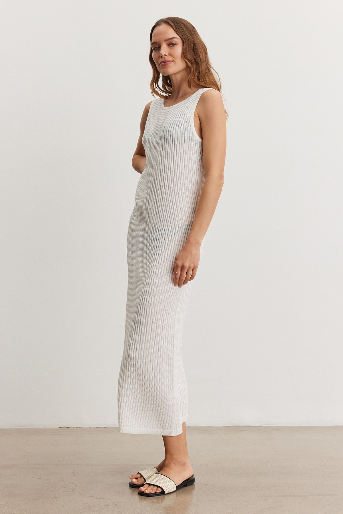 A woman in a white ribbed CALLEN DRESS by Velvet by Graham & Spencer standing in a neutral-toned room, posing with one hand on her hip and wearing black sandals.-36909558956225