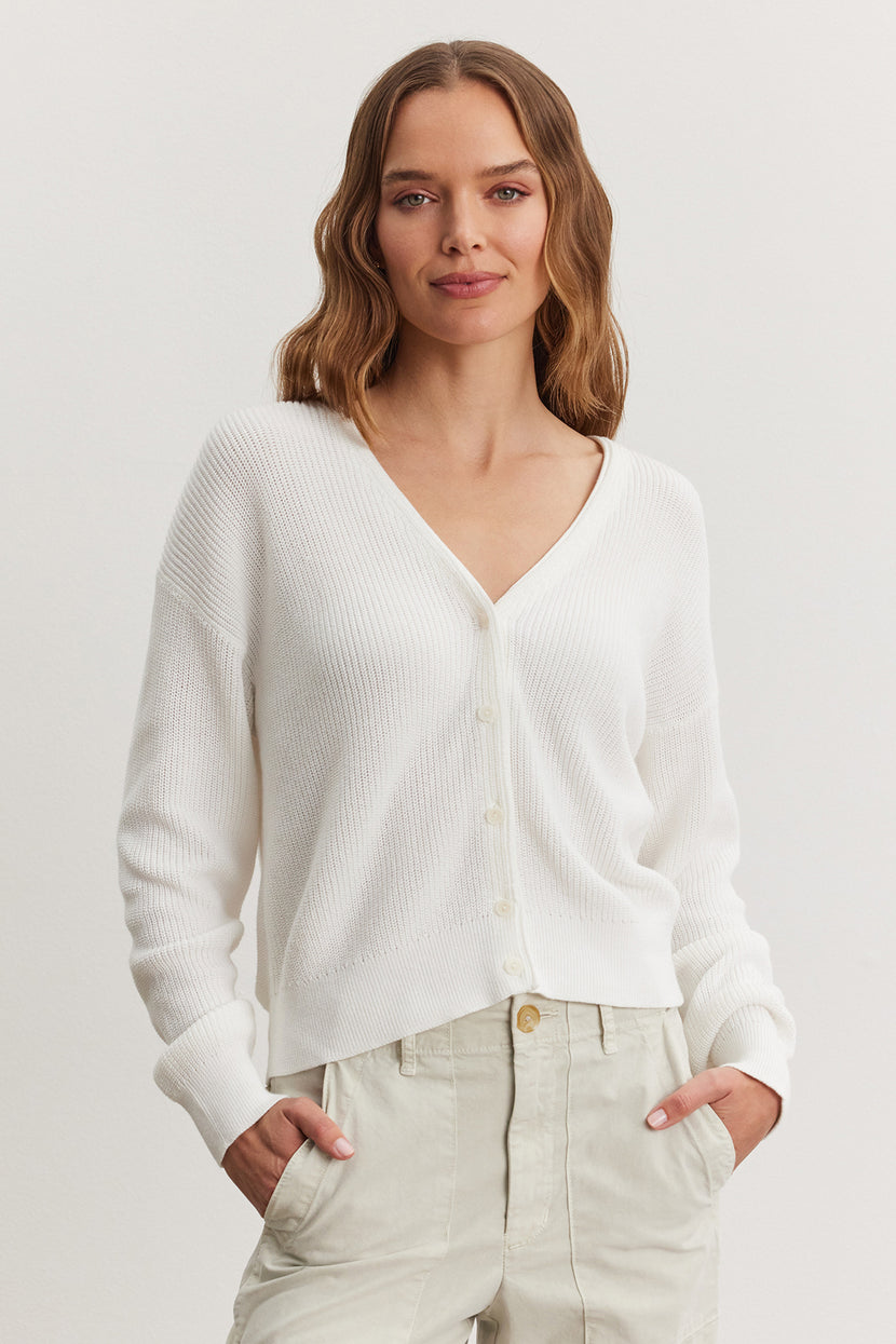 A woman with shoulder-length brown hair wearing a white TAVA Cardigan by Velvet by Graham & Spencer and beige trousers, standing against a plain background.