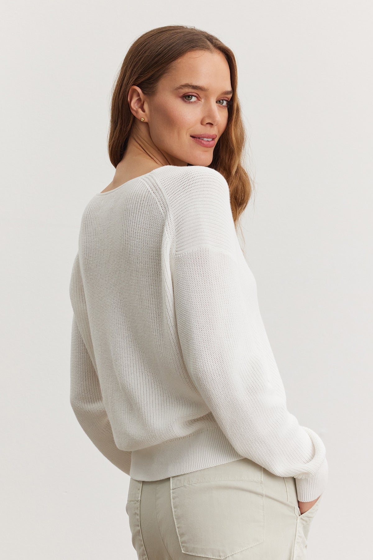   A woman in a white ribbed Velvet by Graham & Spencer TAVA cardigan and light beige pants, standing with her back slightly turned, looking over her shoulder with a soft smile. 