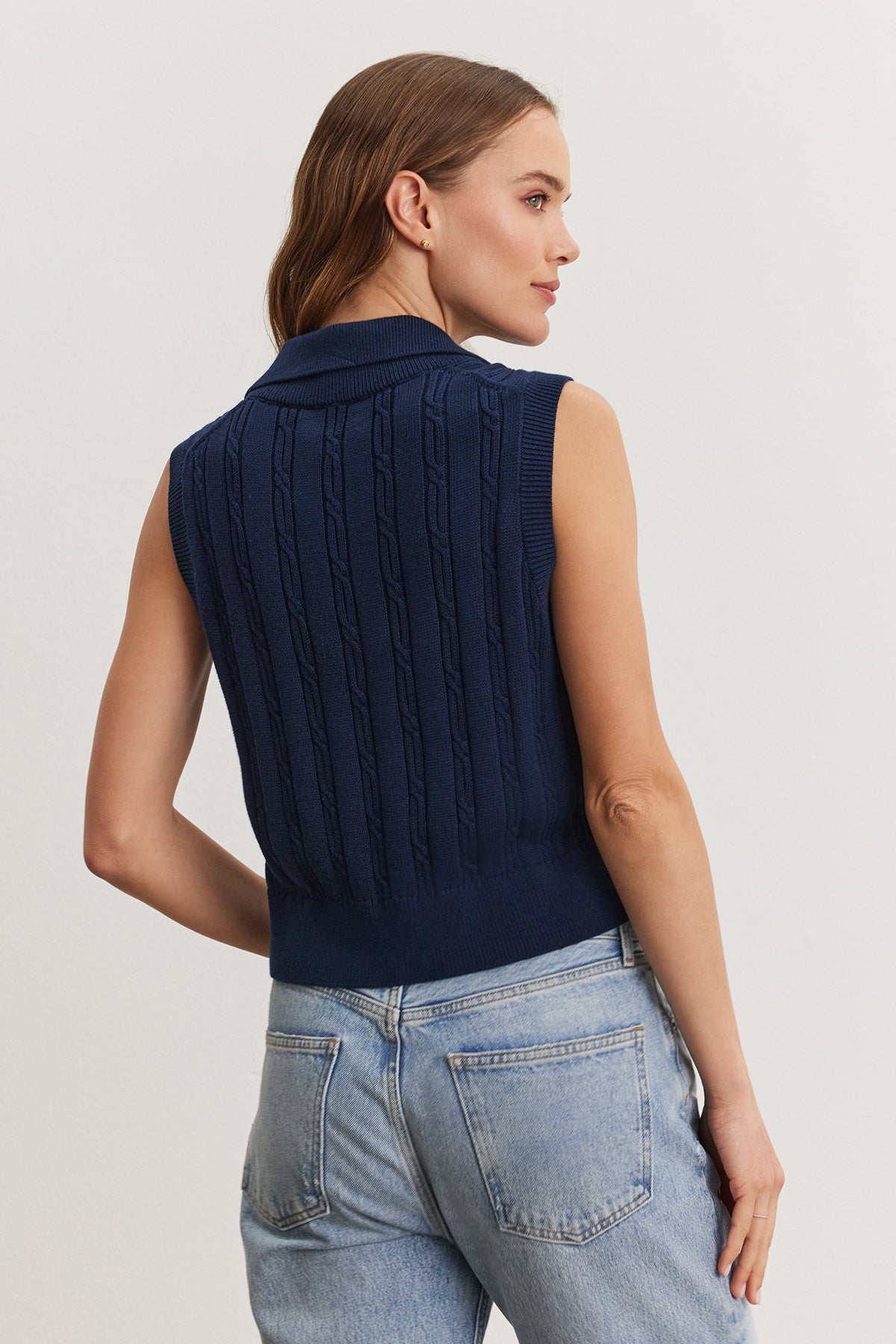 Woman standing facing away from the camera, wearing a blue Velvet by Graham & Spencer Wendy Sweater Vest and light blue jeans, focusing on the sweater's cable knit design.-36909568524481