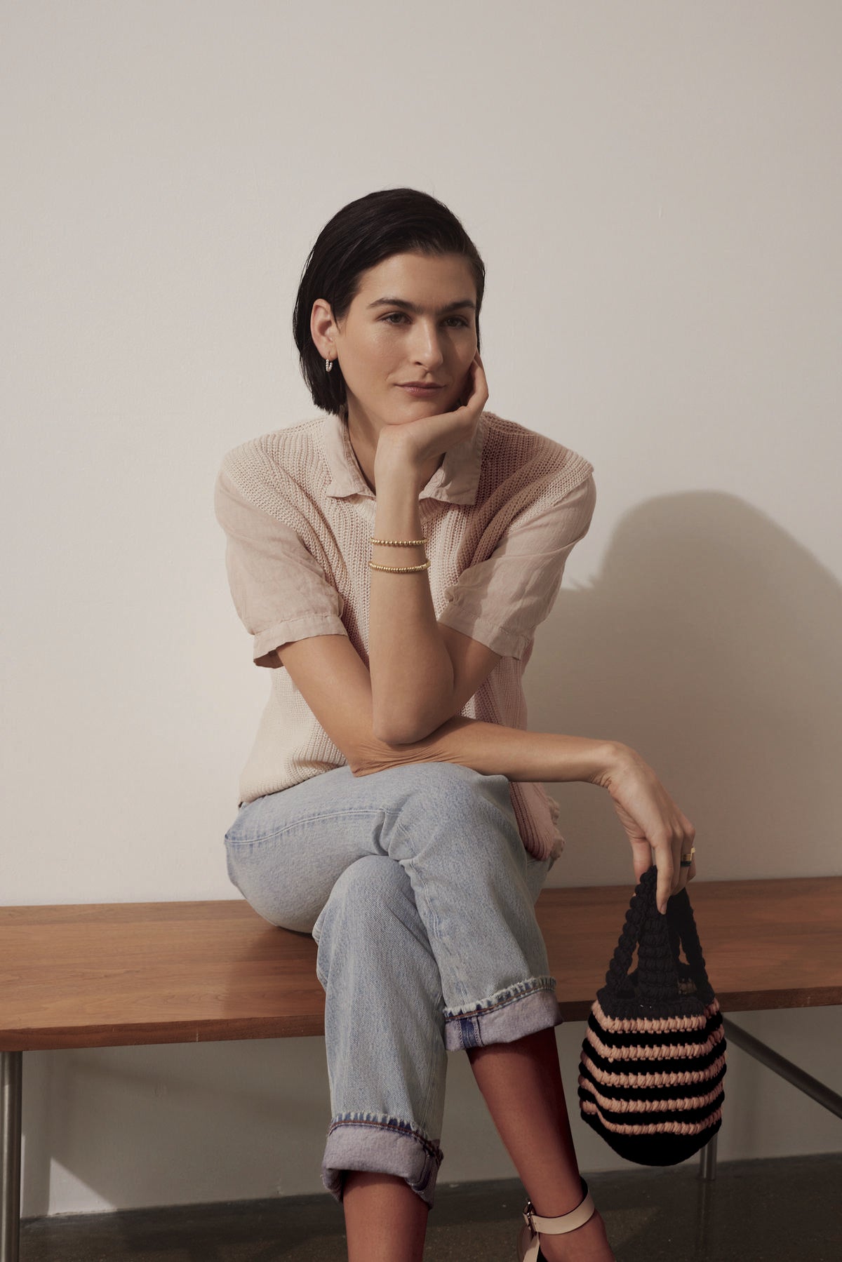   A woman sits on a wooden bench, resting her chin on her hand, wearing a pink blouse, jeans, and holding a Velvet by Jenny Graham KNOX BAG. 