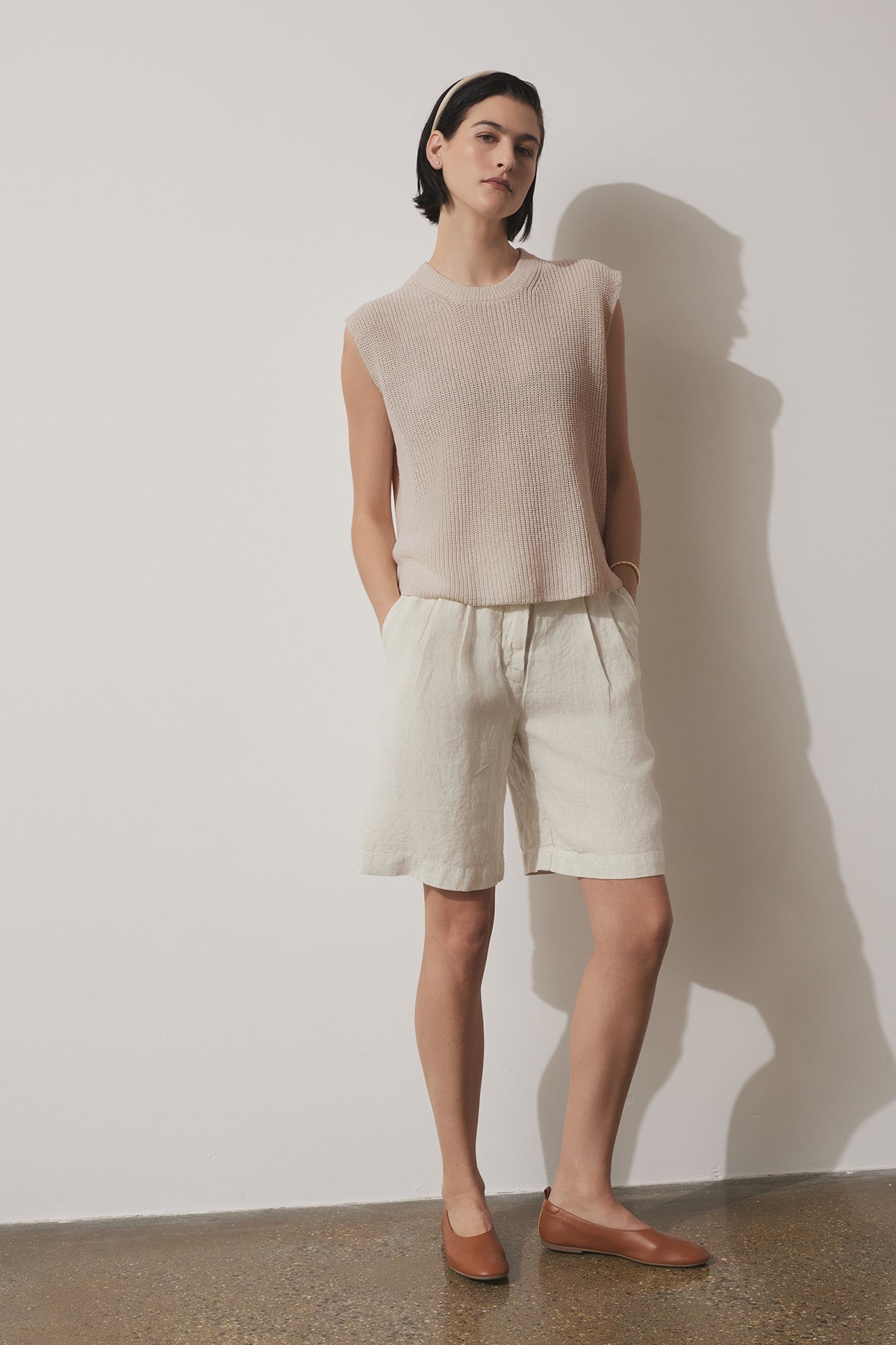 A person stands against a white wall wearing a sleeveless beige top, light-colored LARCHMONT HEAVY LINEN SHORT by Velvet by Jenny Graham, a beige hairband, and brown flat shoes. Hands are in pockets and legs are slightly apart—perfect warm-weather styling.-37629305127105