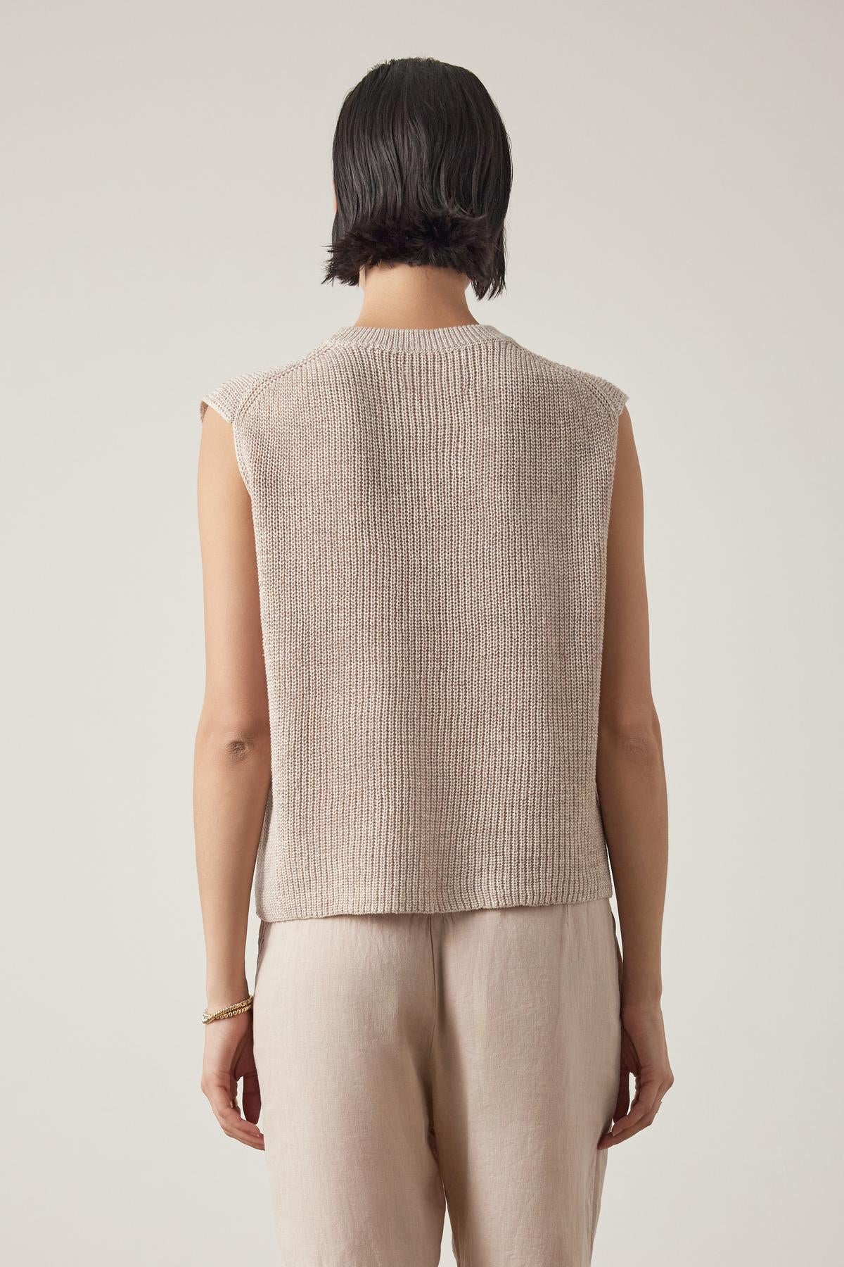   Rear view of a woman wearing a beige GARDENA LINEN SWEATER VEST sleeveless knit top and light linen fabric trousers, standing against a plain background by Velvet by Jenny Graham. 