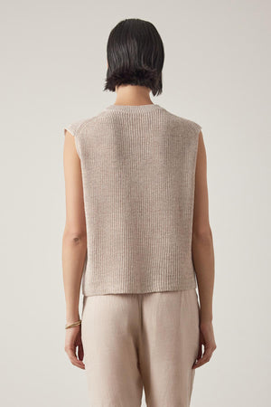 Rear view of a woman wearing a beige GARDENA LINEN SWEATER VEST sleeveless knit top and light linen fabric trousers, standing against a plain background by Velvet by Jenny Graham.