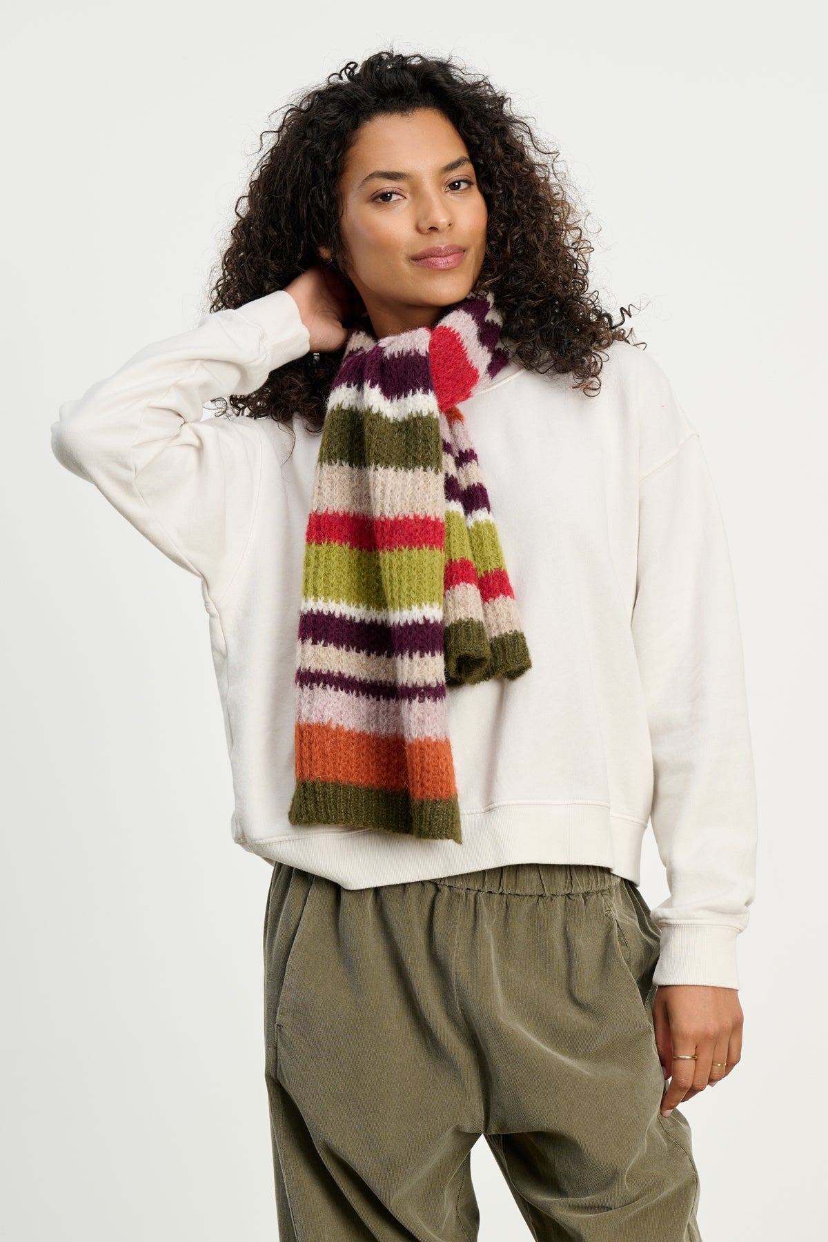 A person wearing an off-white sweatshirt, olive green pants, and the LUCA ALPACA SCARF by Velvet by Graham & Spencer, featuring colorful stripes, stands against a plain light background.-39742257856705