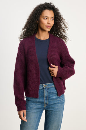A woman wears the RANYA ALPACA CARDIGAN by Velvet by Graham & Spencer over a navy blue t-shirt and blue jeans, standing against a plain light background.