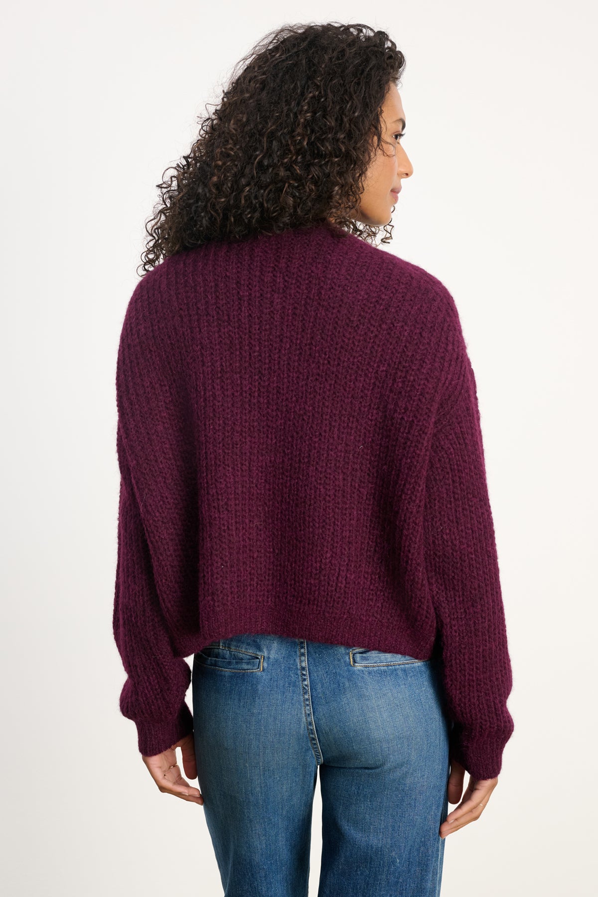 A woman with curly hair wears the RANYA ALPACA CARDIGAN by Velvet by Graham & Spencer in textured dark purple, styled with blue jeans and seen from the back against a plain white background.-40227142729921