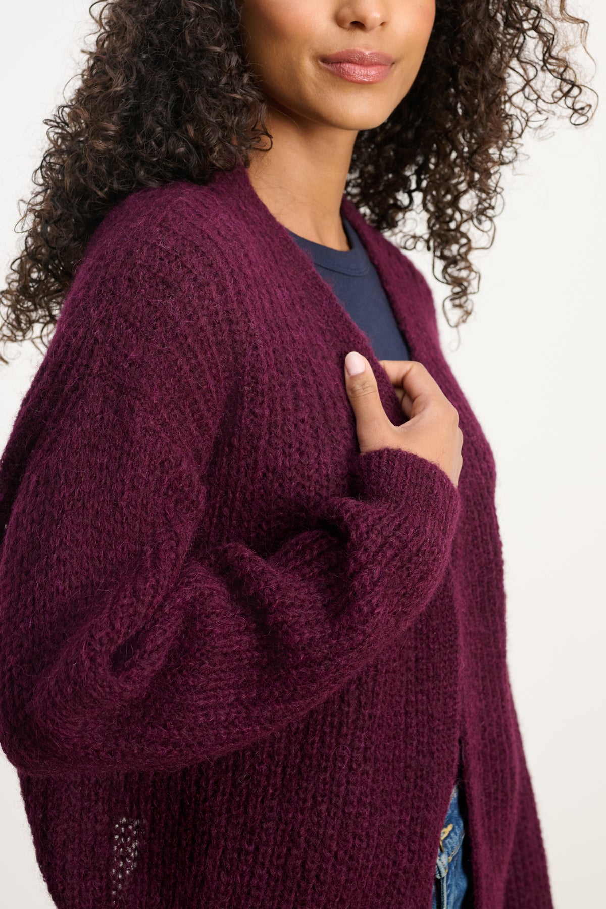 A woman with curly hair wears the RANYA ALPACA CARDIGAN by Velvet by Graham & Spencer in textured dark purple over a navy top, standing against a neutral background.