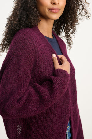 A woman with curly hair wears the RANYA ALPACA CARDIGAN by Velvet by Graham & Spencer in textured dark purple over a navy top, standing against a neutral background.